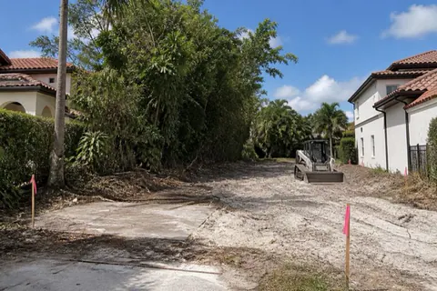 Partially cleared infill homesite in east Boca Raton with pale sand exposed, surveyor stakes set, and overgrown ficus still standing on the uncleared half